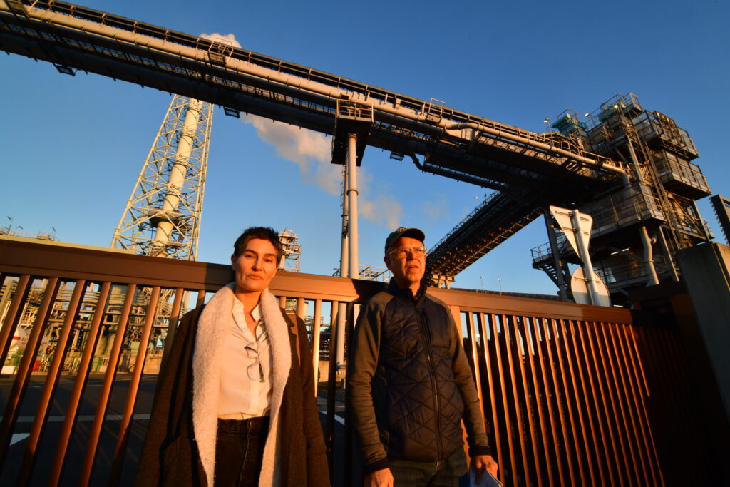 Two people stand in front of a pellet plant in Japan. The sky is blue. Industrial conveyors run laterally high above. A smoke stack can be seen as well.