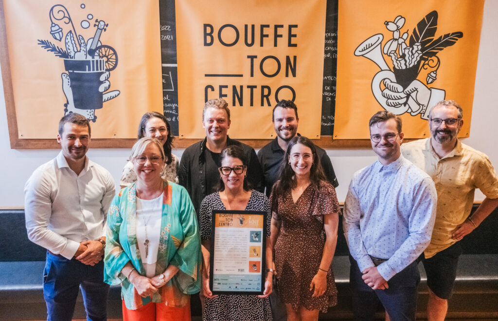 Nine people stand in front of an orange and black banner which reads "Bouffe ton centro". The person in the centre is holding up a framed certificate.