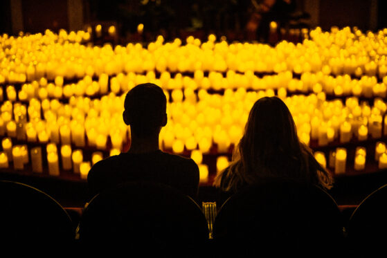 Two people in the foreground sit with their backs to the camera. They are in silhouette as hundreds of candles can be seen in the background. s