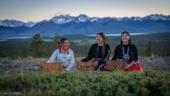 Three Tŝilhqot’in women, dressed in a mix of traditional and modern attire, sit smiling in a field holding qatŝ’ay (coiled root baskets), with the sun seting over distant mountains in the background