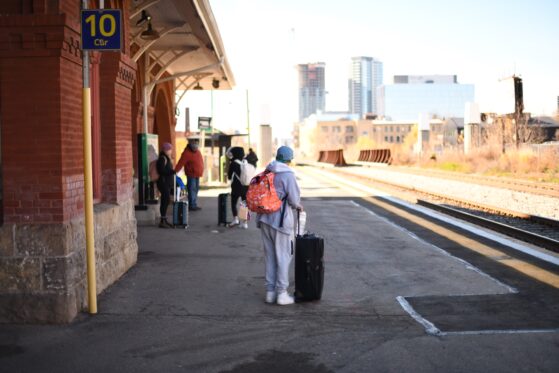 A person wearing a grey track suit stands with their rolling suitcase while waiting for a train on a train station platform.